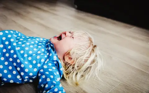 Mała blondynka w niebieskiej sukience w kropki leży na podłodze, krzycząc. To klasyczny bunt 2 latka objawy.