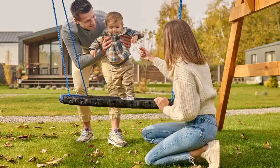 Radosne zabawy w ogrodzie: tata podtrzymuje dziecko na huśtawce, mama zachęca do zabawy.