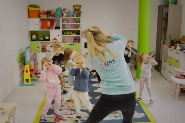 Dzieci na zajęciach tańca wykonują proste układy taneczne do znanych piosenek, naśladując ruchy nauczycielki.
